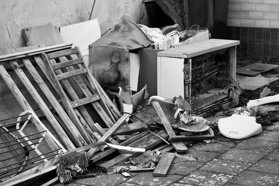 A pile of black rubbish bags and discarded packaging materials, including cardboard and plastic, stacked against a textured beige wall beneath a red metal fire door covered in graffiti and stickers. T
