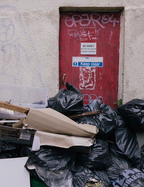 A middle-aged woman with short dark hair, wearing a black T-shirt with white and grey text on the front, is seen disposing of waste into a stainless steel trash bin on a paved outdoor walkway. She is 