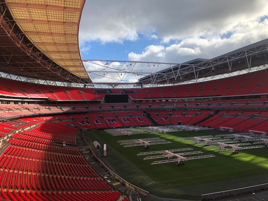A panoramic view of a large, empty football stadium with a bright green grass pitch marked with white lines for gameplay. The stadium features multiple tiered seating areas primarily in red, with sect