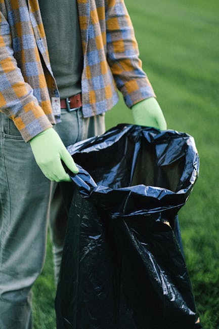 A large pile of mixed waste and rubbish bags situated on a paved sidewalk in an urban area. The refuse includes cardboard boxes, plastic bags, and various discarded packaging materials, some partially